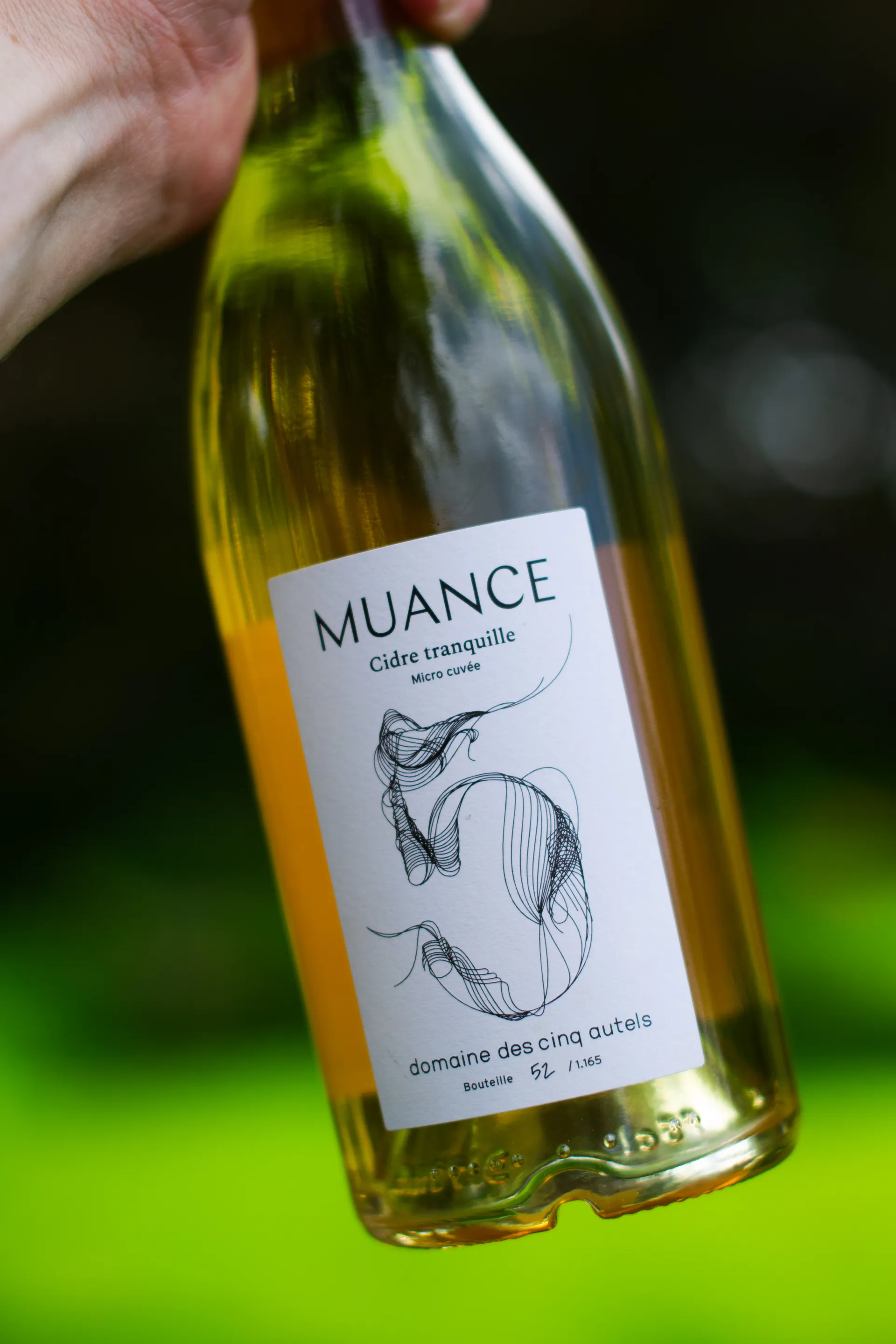Bouteille de cidre Muance tenue à la main en plein air, étiquette aux pommes stylisées visible